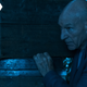 Jean-Luc Picard, in the dark, hides behind a stack of barrels and boxes, resting one hand on the pile.